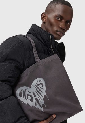 Borsa tote grigia in tela con un grande design grafico a forma di cuore argentato, caratterizzata da testo stilizzato e gocce, tenuta da una persona con una giacca nera.