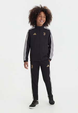 Barn med krøllet hår, som smiler, iført sort Adidas Juventus træningsdragt med hvide striber og guldlogoer, stående med afslappede hænder.