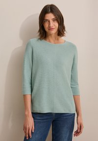 Top in maglia verde chiaro, con tessuto testurizzato, scollatura rotonda e maniche a tre quarti, abbinato a jeans in denim blu. Taglio semplice e rilassato.