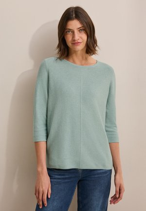 Top en tricot texturé vert clair avec un col rond et des manches trois-quarts, associé à un jean en denim bleu. Coupe simple et décontractée.