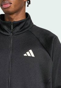 Casaco atlético preto com gola alta e fecho de zíper. Apresenta um logo da adidas branco na área do peito esquerdo. Textura suave.