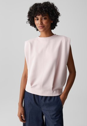 Frau mit lockigem Haar, die ein hellrosa ärmelloses Oberteil mit Schulterpolstern und dunkelblaue Hosen trägt, steht mit einer Hand in der Tasche.