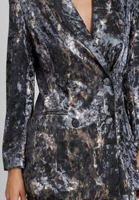 Blazer de doble botonadura en negro con patrones abstractos en gris y marrón. Textura aterciopelada, dos botones grandes y cuello de muesca.