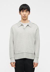 Νεαρός με κοντά μαύρα μαλλιά, φορώντας ανοιχτό γκρι πλεκτό πουλόβερ με γιακά πάνω από λευκό πουκάμισο και μαύρο παντελόνι, στέκεται μπροστά σε λευκό φόντο.