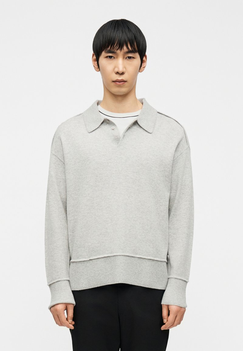 Νεαρός με κοντά μαύρα μαλλιά, φορώντας ανοιχτό γκρι πλεκτό πουλόβερ με γιακά πάνω από λευκό πουκάμισο και μαύρο παντελόνι, στέκεται μπροστά σε λευκό φόντο.