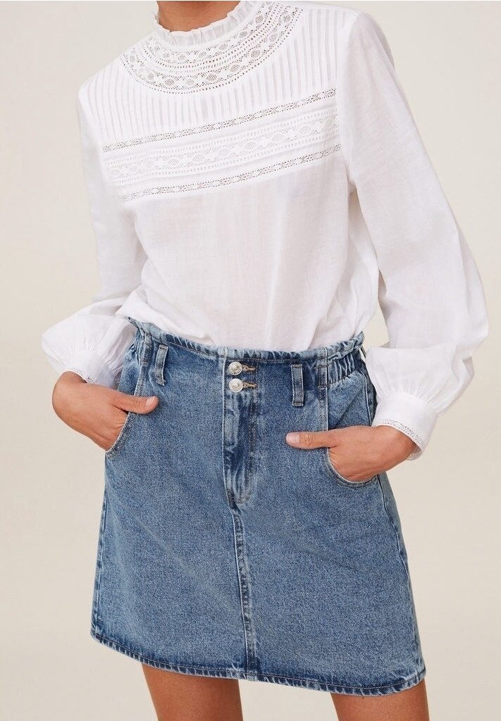 Blouse blanche manches longues avec dentelle et plis, associée à une jupe en jean bleu taille haute avec poches avant et double bouton.