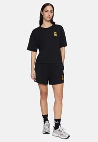 T-shirt nera in cotone a maniche corte con un logo giallo sul petto, abbinata a pantaloni corti neri e scarpe da atletica argento.
