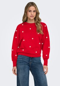 Red sweatshirt with white floral embroidery, round neckline, and long, puffed sleeves. Paired with denim jeans, casual fit.