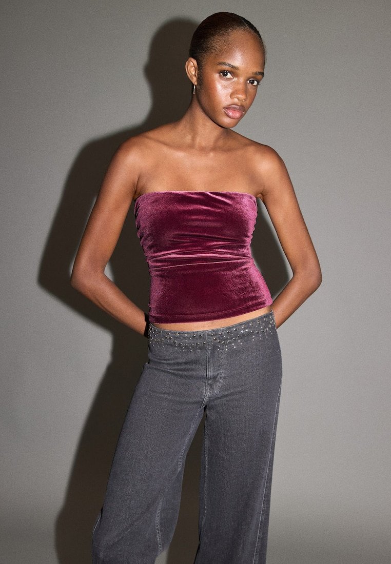 Top z vínového sametu bez ramínek s přiléhavým střihem, doplněný tmavě šedými džínami s ozdobnými hroty na pasu.