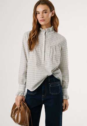 White and gray checked shirt with a ruffled collar and sleeves, featuring a button placket and a breast pocket, paired with dark jeans and a brown bag.