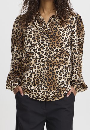 Femme portant un chemisier à manches longues imprimé léopard avec un col à volants et un pantalon noir, mains dans les poches, sur un fond uni.