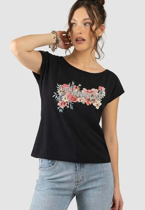 Giovane donna con capelli ricci indossa una t-shirt nera con stampa floreale e jeans azzurri decorati con strass, posando con una mano vicino alla testa.