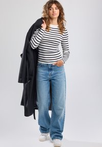 Striped, ribbed long-sleeve top in black and white; loose-fit light blue denim jeans; black coat draped over shoulder; white sneakers.