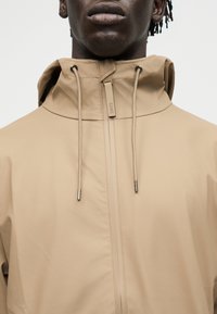 Nahaufnahme einer Person, die eine beige Kapuzenregenjacke mit Kordelzug und einem zentralen Reißverschluss mit der Marke "RAINS" trägt.