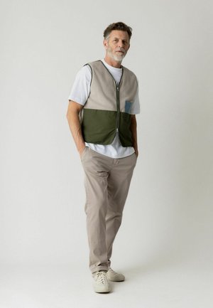 Middelaldrende mand stående med hænderne i lommerne iført beige bukser, hvide sneakers, hvid T-shirt og en tofarvet vest med lynlås.