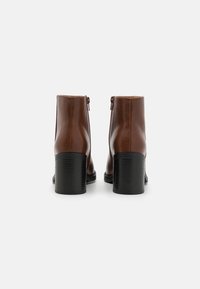 Botas de cuero marrón hasta el tobillo con un acabado suave, cierre de cremallera trasero y un tacón bloque negro estriado. Diseño minimalista con una silueta elegante.