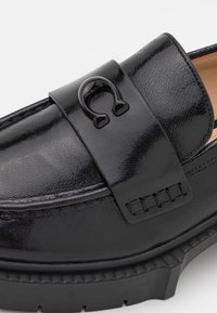 Gros plan d'un mocassin en cuir noir avec une semelle épaisse et un détail décoratif métallique noir en forme de fer à cheval sur la bride.