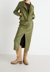 Femme portant une veste en cuir olive et une jupe fendue assortie avec des bottes en daim beige jusqu'au genou, se tenant devant un fond blanc uni.