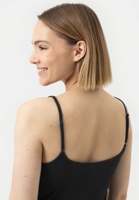 Młoda kobieta z krótkimi blond włosami, ubrana w czarny top na ramiączkach, uśmiechająca się i patrząca w lewo na tle białego tła.