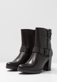 Botas de tobillo de piel negra con un tacón grueso, cremallera lateral y una cinta ancha con detalle de hebilla. Textura suave y punta redondeada.