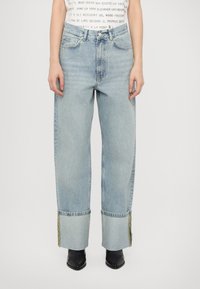 Jeans de denim azul claro com um corte largo, bainhas desfiadas e costura amarela em contraste. Apresenta bolsos frontais e um design clássico de cinco bolsos.