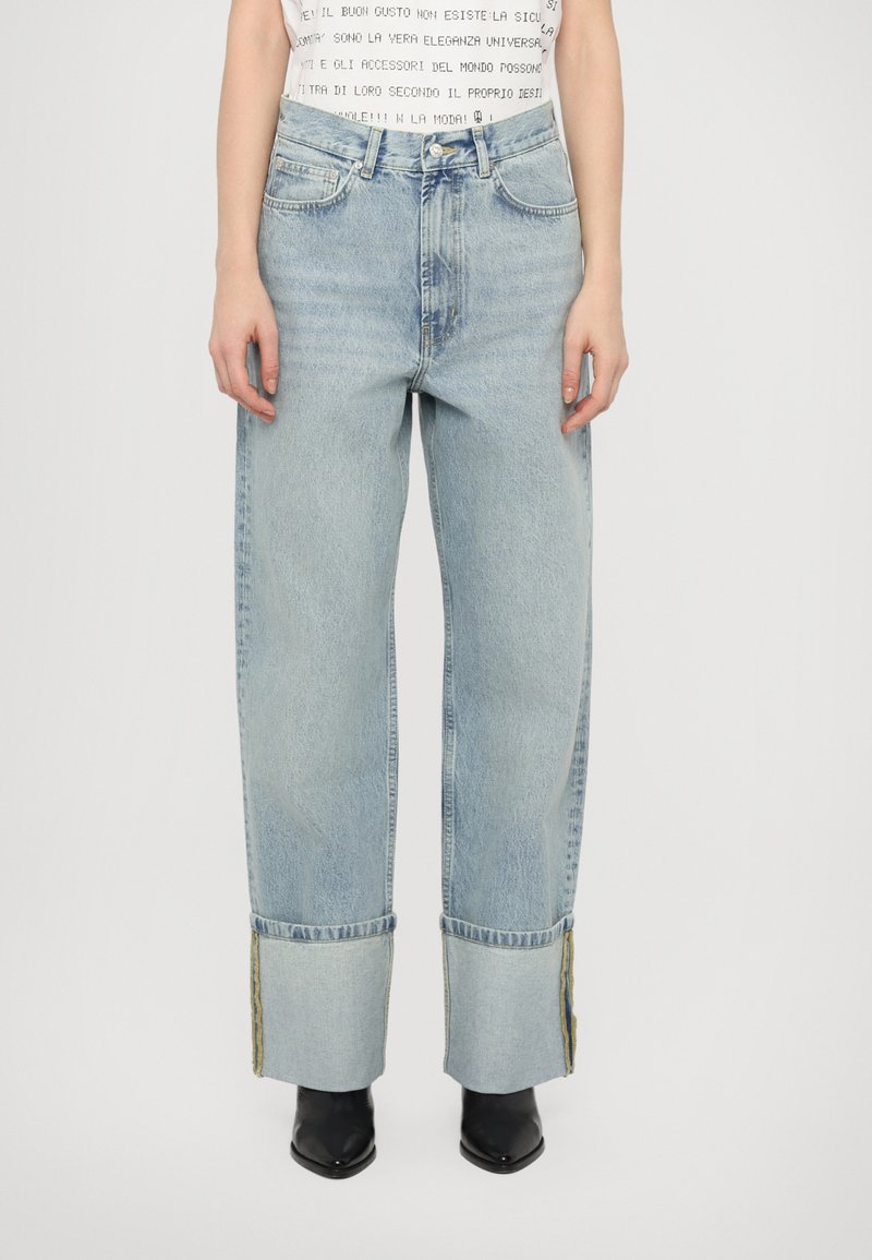 Jeans de denim azul claro com um corte largo, bainhas desfiadas e costura amarela em contraste. Apresenta bolsos frontais e um design clássico de cinco bolsos.