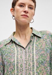 Το φλοράλ μπλουζάκι έχει πράσινο φόντο με ροζ, μπλε και άσπρα σχέδια. Με κουμπιά μπροστά και γιακά, υφασμάτινη υφή και διακοσμητικά στοιχεία.