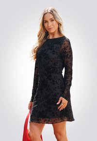Vestido negro de mangas largas con encaje floral superpuesto y dobladillo con forma de ola, acompañado de un bolso rojo. Tejido suave, silueta ajustada.