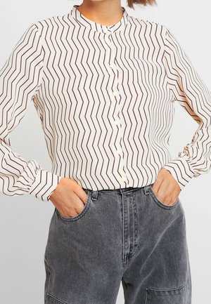 Personne portant une chemise blanche à boutons avec des rayures en zigzag marron et un jean noir délavé taille haute avec les mains dans les poches.