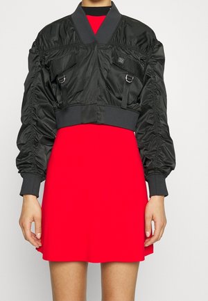 Oříznutá černá bomber bunda s nařasenými rukávy a předními kapsami přes jasně červené krátké šaty, zobrazená od krku po polovinu stehen.