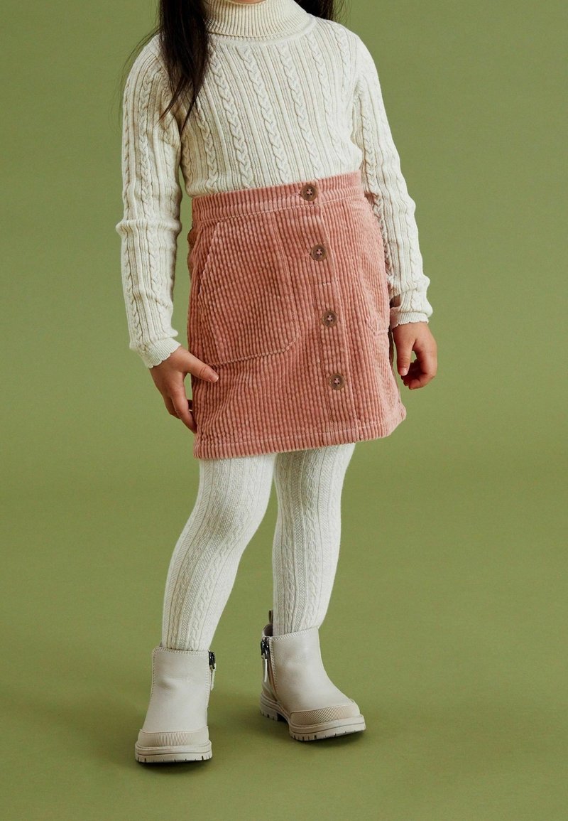 Enfant portant un col roulé en maille torsadée blanche, des collants blancs à motifs, une jupe boutonnée en velours côtelé rose et des bottines beige sur fond vert.