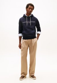 Sudadera con capucha azul marino con cordones y logo blancos, combinada con pantalones beige. El modelo está de pie con las manos a los lados, vistiendo zapatillas marrones.