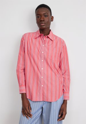 Woman wearing loose red and white striped button-up shirt with light blue pinstriped pants standing against white background.