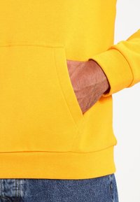 Felpa gialla con tasca kangaroo. Presenta una texture morbida e orli a coste, progettata per un abbigliamento casual. Dettagli di cucitura evidenti all'orlo.