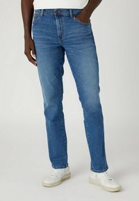 Vaqueros de mezclilla azul con un diseño de pierna recta, sutil desvanecimiento y estilo clásico de cinco bolsillos. Llevados con zapatillas blancas.