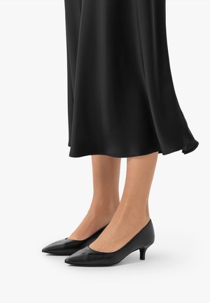 Nogi w czarnych, spiczastych butach na niskim obcasie typu kitten heel oraz w czarnej, zwiewnej spódnicy na białym tle.