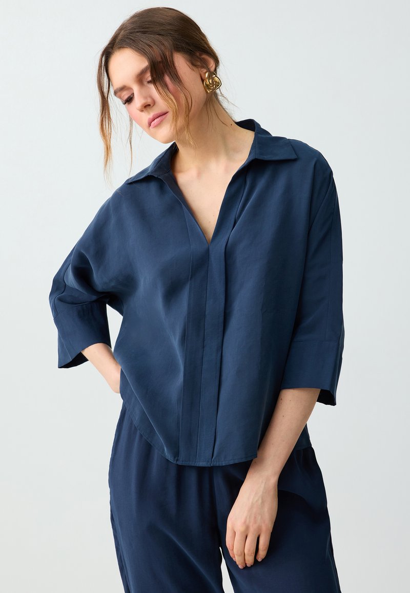 Femme en chemise bleu marine ample avec un large col et un pantalon assorti, portant des boucles d’oreilles en anneau torsadé doré, tête inclinée vers le bas, yeux fermés.