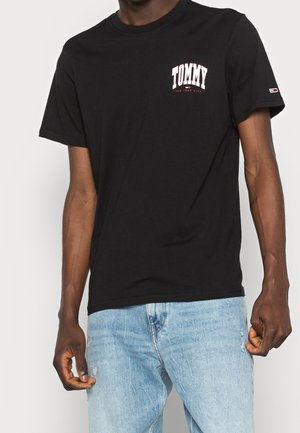 Black cotton t-shirt featuring red and white "TOMMY" logo on the left chest. Short sleeves and crew neck design. Worn with light blue denim jeans.