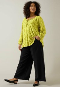 Lysgul texturerad blus med rundad halsringning och rynkade ärmar, i kombination med löst sittande svarta byxor och svarta slip-on skor.