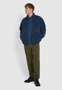 Giacca in pile blu con pannello grigio scuro, colletto alto e chiusura con zip, abbinata a una camicia blu e pantaloni verde oliva, indossata con scarpe nere.