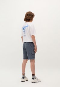 Camicetta bianca a maniche corte con testo grafico blu sul retro, abbinata a pantaloni cargo grigi e scarpe da ginnastica bianche con dettagli blu navy.