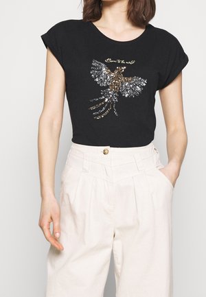 Kvinde iført sort T-shirt med op-rullede ærmer, der har en paliet-beklædt fugl og teksten "Born to be wild", kombineret med højtaljede hvide bukser.