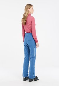Pantaloni da sci blu con vita alta, tasche laterali con zip e polsini per stivali regolabili, abbinati a una maglietta a maniche lunghe rosa.