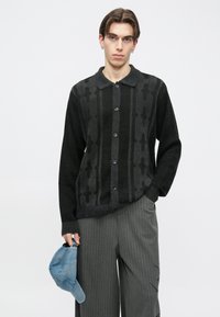 Džemper na kopčanje pleteni u crnoj i sivoj boji s vertikalnim uzorcima. Ima ovratnik i duge rukave. Uz to dolazi plava kapa.