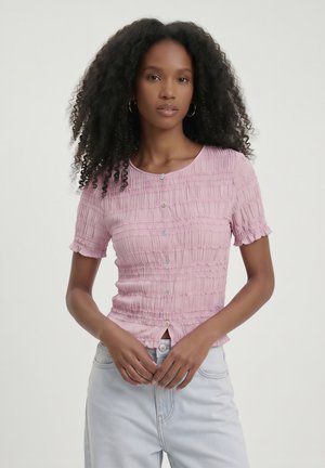 Kvinde med krøllet sort hår iført en lyserød rynket knapkortærmet top og lyseblå jeans, stående mod en ensfarvet baggrund.