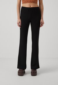 Black flared pants with an elastic waistband, crafted from textured fabric, paired with brown platform clogs on a neutral background.