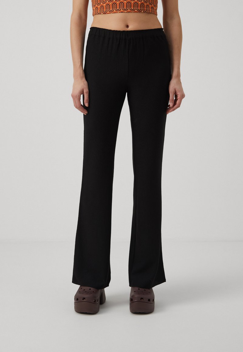Black flared pants with an elastic waistband, crafted from textured fabric, paired with brown platform clogs on a neutral background.
