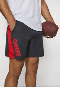 Shorts de sport gris avec un panneau latéral rouge portant le mot "CHIEFS" en lettres majuscules. Tissu léger, deux poches latérales et coupe flexible.