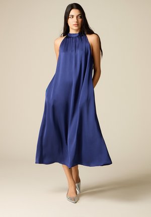 Frau in einem ärmellosen, locker sitzenden, königsblauen Midikleid und silbernen spitzen High Heels, vor einem schlichten beige-farbenen Hintergrund stehend.