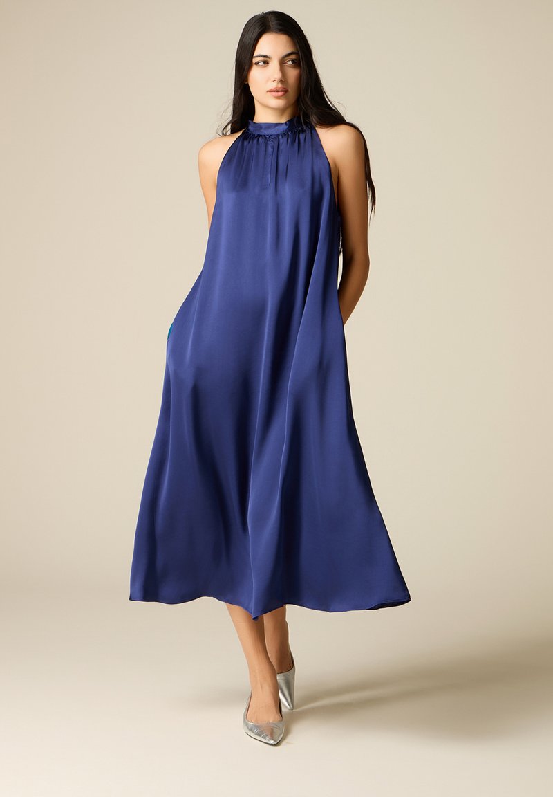 Femeie într-o rochie midi largă, fără mâneci, de culoare albastru regal și pantofi cu toc ascuțit argintii, stând în fața unui fundal bej simplu.